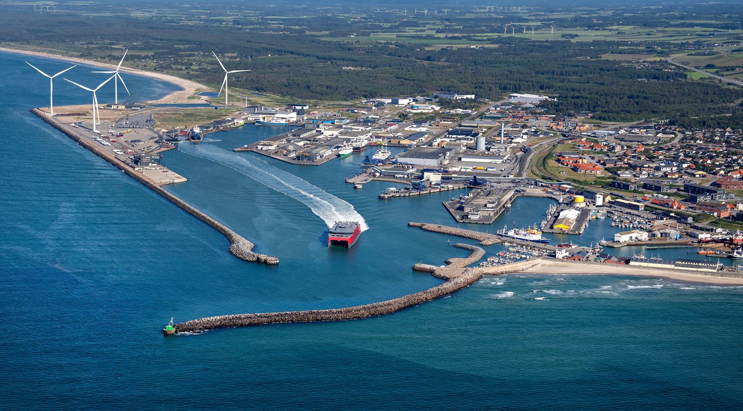 Foto fra luften af Hirtshals Havn, havet i forgrunden, havnen hvor en færge sejler ud, vindmøller på havnen, byen og landskab i baggrunden med træer og vindmøller